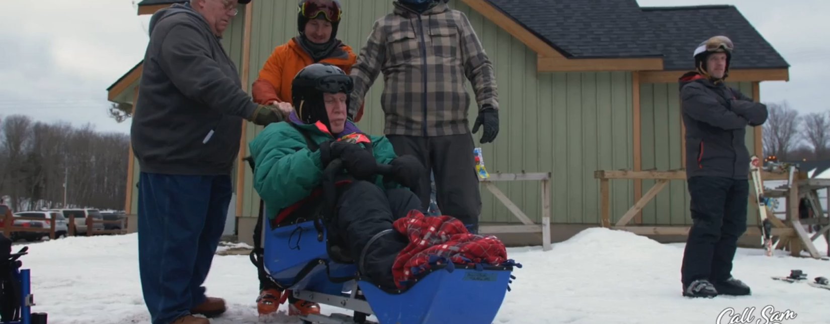 Three people standing behind someone in a sled made for disabled people on a snowy mountain side in front of a buidling