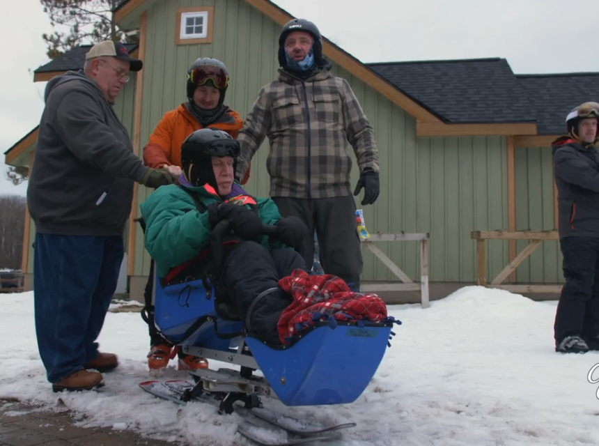 Three people standing behind someone in a sled made for disabled people on a snowy mountain side in front of a buidling